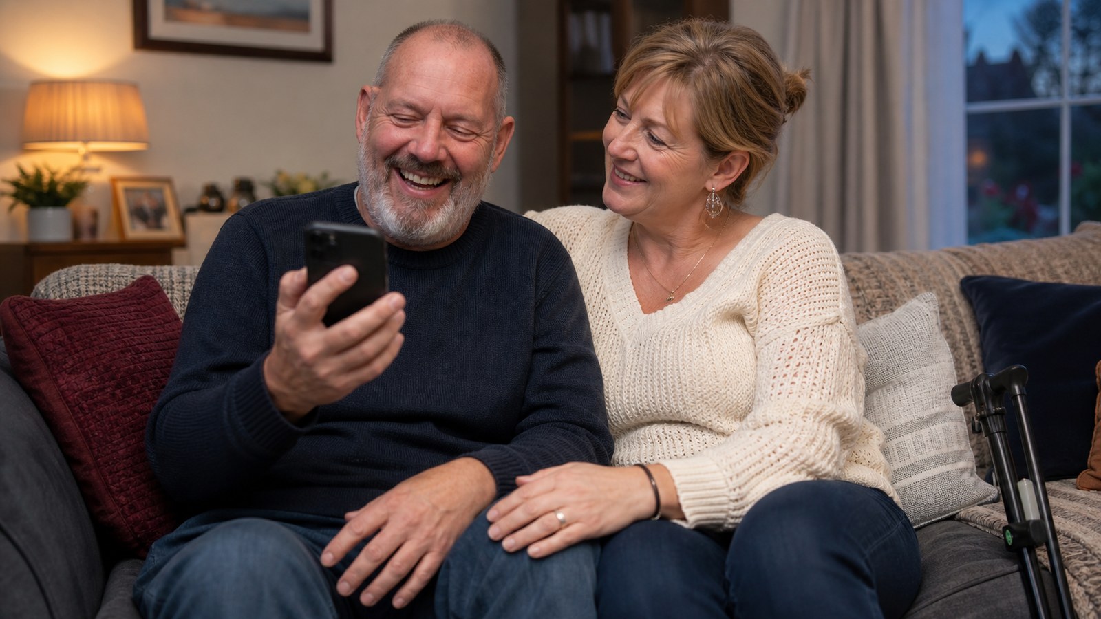 A couple laughing together over a smartphone at home, with a walking stick beside the sofa
